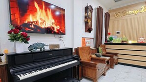 A living room with a piano, a television displaying a fireplace, and a reception desk. - Hostel Nha Tho (Lam Vien - Da Lat)
