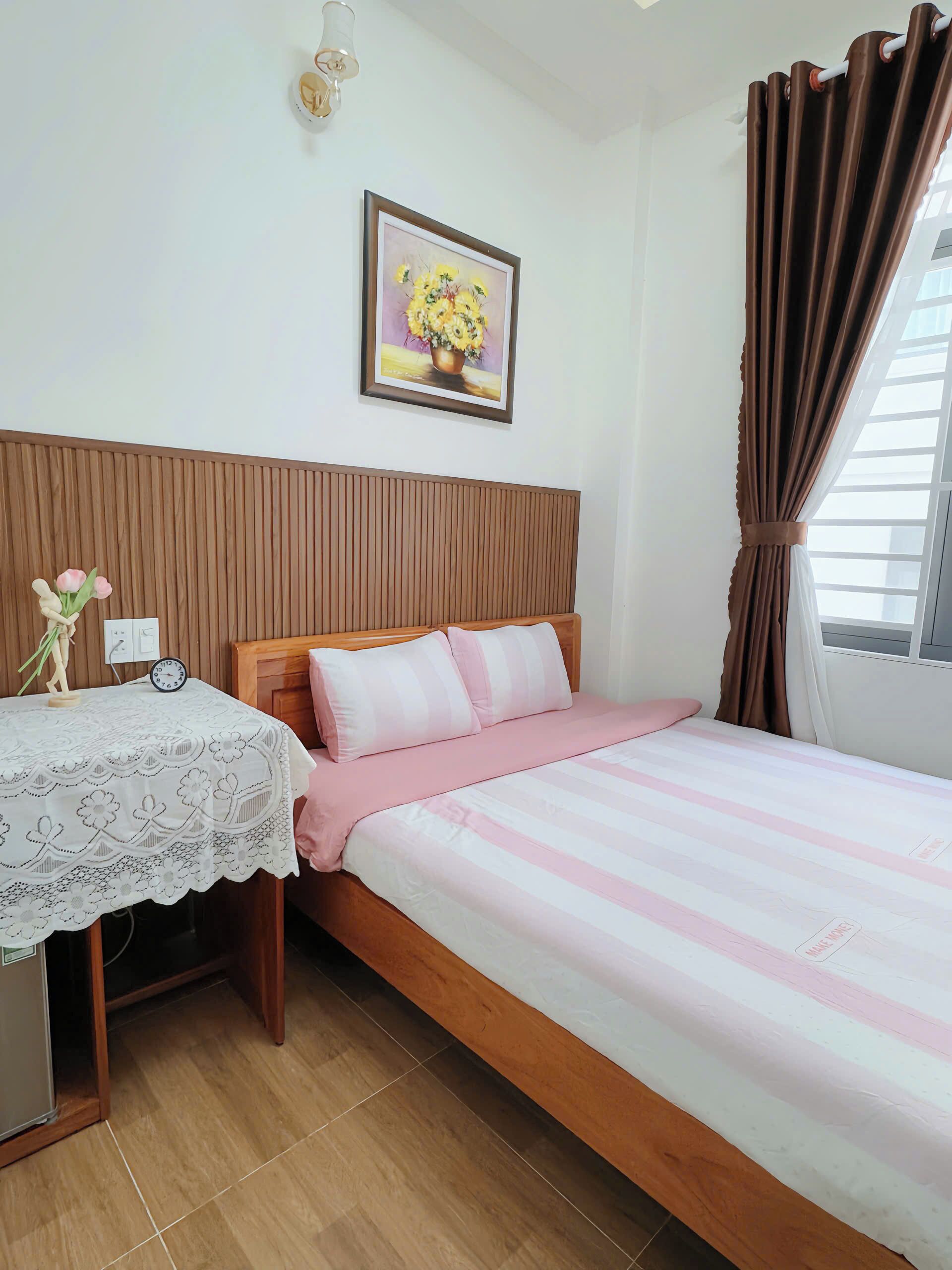 A neatly arranged bedroom with a wooden headboard, a bed with pink and white bedding, a bedside table with a lace tablecloth, a clock, and a framed floral painting on the wall.
