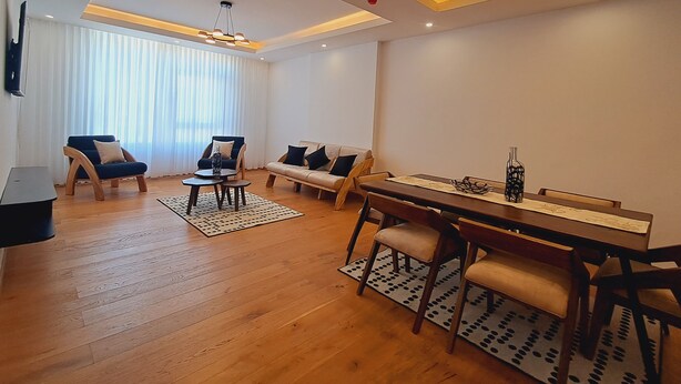 Una sala de estar moderna con piso de madera, un conjunto de sofás, una mesa de comedor con sillas y un televisor montado en la pared.
