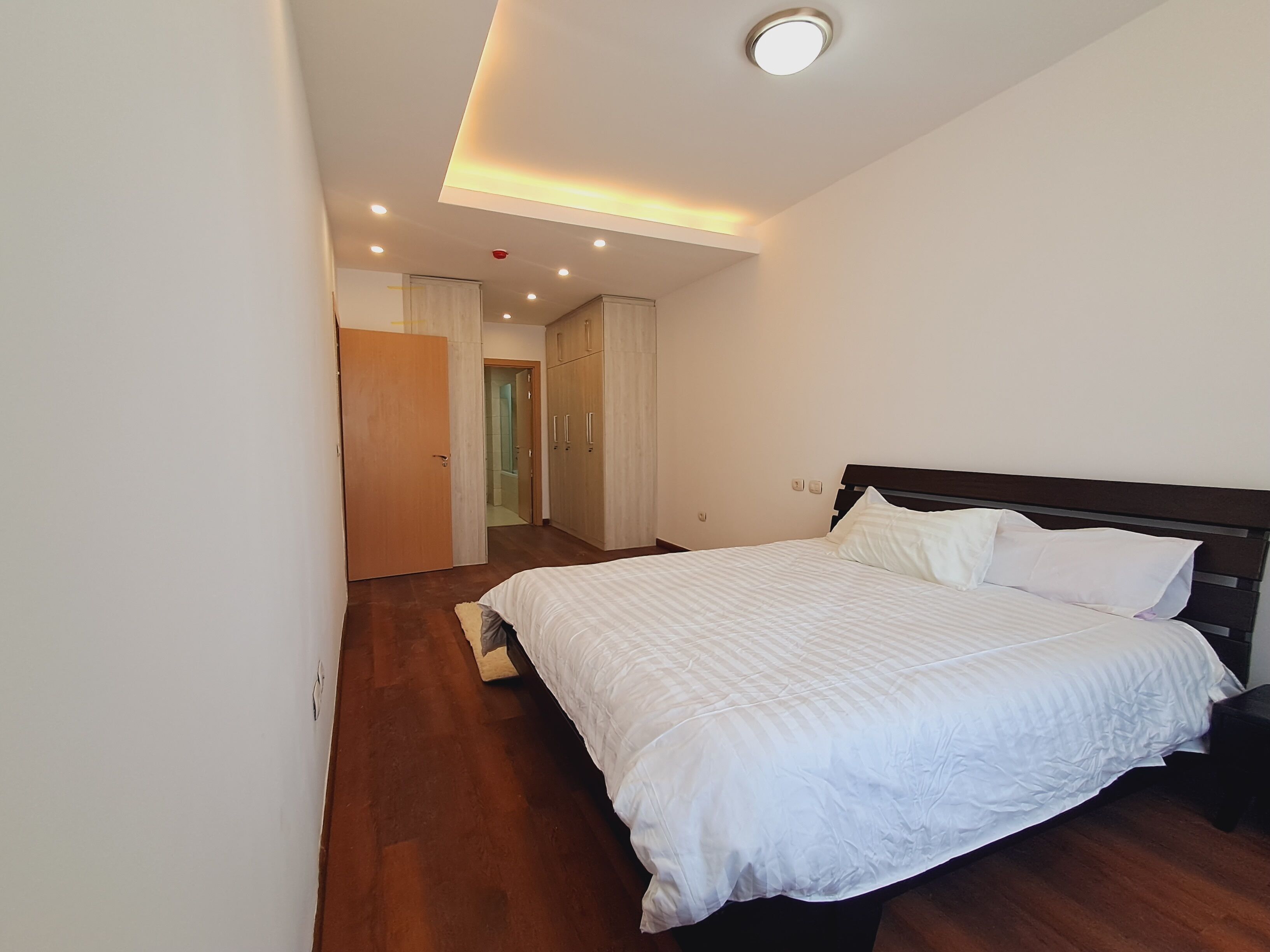 A bedroom with a large bed, wooden flooring, and a ceiling light fixture.