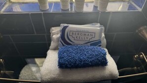 Bany amb un espai de banyera de vidre, una tovallola blanca, una tovallola blava i diversos productes de dutxa Bergman Kelly a l'estant.