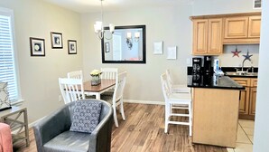 A compact kitchen with wooden cabinets, a central island, and a dining area with white chairs and a table. - Sunchase 204 | Pool & Hot Tub | Steps to the Beach (Gulf Shores)