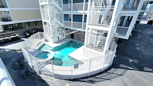 A rooftop pool with a hot tub, surrounded by a white metal fence, and a multi-level building in the background. - Sunchase 204 | Pool & Hot Tub | Steps to the Beach (Gulf Shores)