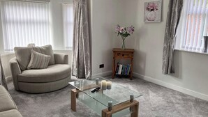 Een woonkamer met een ronde beige bank, een glazen salontafel, een klein houten bijzettafeltje met boeken en een vaas met bloemen.