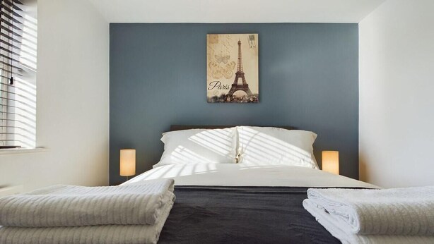A neatly made bed with a dark headboard, white pillows, and a folded white towel on each side. A framed picture of the Eiffel Tower is hung above the bed. - Sleeps6- FamilyFriendly- FreeParking- 3Br (Stafford)