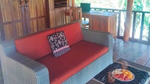 Un sofá de mimbre con un cojín rojo, una mesita con una bandeja de frutas y una estructura de techo de paja.