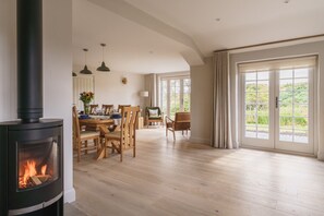 A spacious living room with a wood-burning stove, a dining area with wooden furniture, and large windows offering a view of greenery.