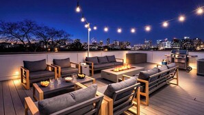 A rooftop patio with outdoor seating, a fire pit, and a grill, overlooking a city skyline at night. - Encore Estates - Rooftop deck & skyline views (Nashville)