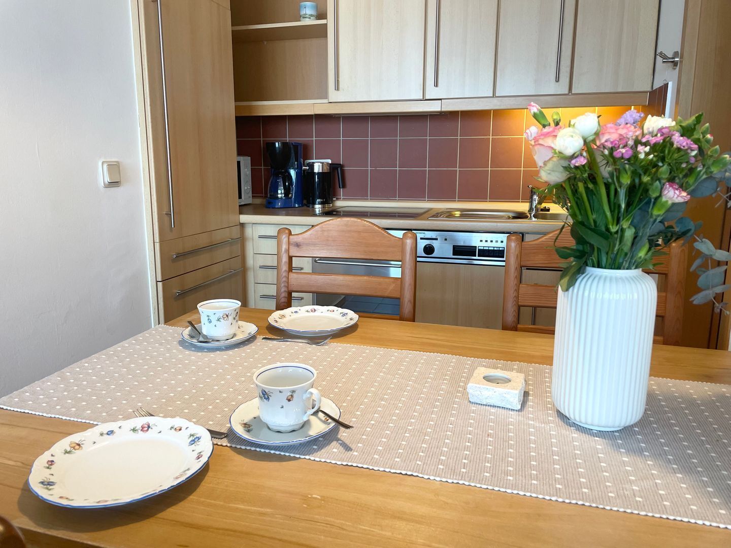 Una cocina con armarios de madera, una cafetera, un lavabo y un florero con flores sobre la mesa.