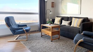 A modern living room with a view of the city, a blue armchair, a dark sofa, and a wooden coffee table. - FB12.06 - Apartment for 2 persons with sea view, swimming pool and sauna - (Cuxhaven)
