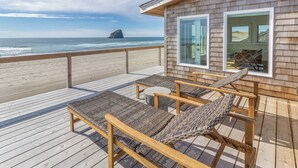 Een houten terras met rieten stoelen en een kleine ronde tafel, met uitzicht op de oceaan en een verre rotsformatie.