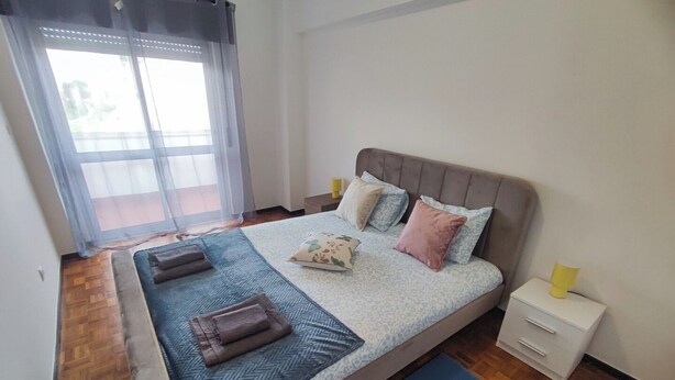A bedroom with a bed, a nightstand, a window with curtains, and a wooden floor. - Lisbon spacious T2 in Damaia, Lisbon (Amadora)