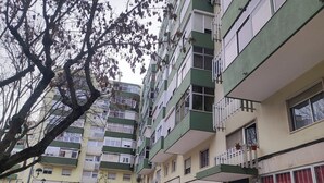 A multi-story residential building with balconies, a small garden area, and various storefronts. - Lisbon spacious T2 in Damaia, Lisbon (Amadora)