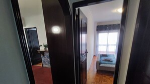 A hallway leading to a bedroom with a bed, a window with curtains, and a small table with a plant. - Lisbon spacious T2 in Damaia, Lisbon (Amadora)