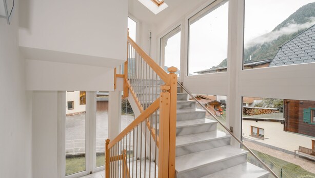 Un escalier moderne avec des rampes en bois et des marches en marbre, menant à un deuxième étage doté de grandes fenêtres offrant une vue sur un paysage montagneux.