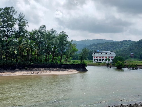A riverside view with a building on the opposite bank, surrounded by lush greenery and hills in the background. - Shobha Farmhouse (Murud)