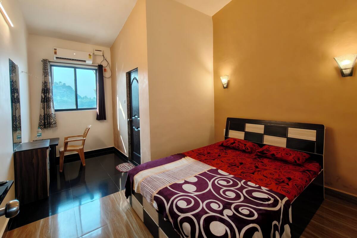 A bedroom with a bed, a desk, and a chair. There is a window with curtains, an air conditioning unit, and a wall-mounted light fixture.