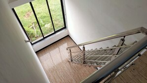 A staircase with a metal handrail and a view of a grassy area outside through a large window. - Shobha Farmhouse (Murud)