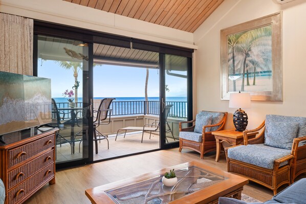 A room with a glass door leading to a balcony with a view of the ocean. There is a wicker sofa, a small table with a lamp, and a television on a stand.