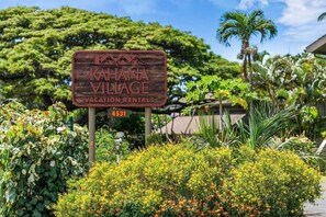 Un letrero de madera indica Kahana Village, con alquileres vacacionales, rodeado de exuberante follaje y palmeras.