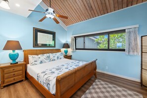 A bedroom with a bed, wooden headboard, two bedside tables with lamps, a ceiling fan, a window with curtains, and a wall-mounted mirror.
