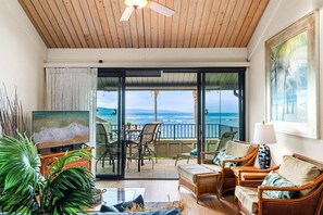 A spacious living room with a large TV, comfortable seating, a balcony with a view of the ocean, and a ceiling fan.