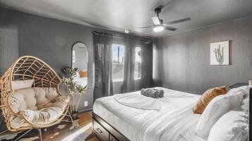 Un dormitorio con cama, ventilador de techo, una silla colgante y una ventana con cortinas.