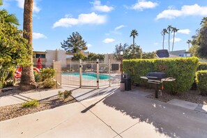 A gated pool area with a barbecue grill, surrounded by palm trees and a hedge.