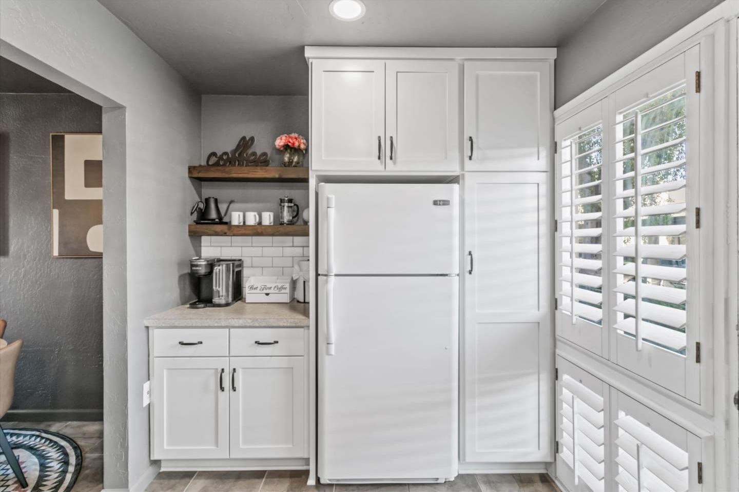 Una cocina con armarios blancos, un refrigerador empotrado, una estación de café y un ventanal con persianas.
