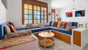 Un salon moderne avec un canapé bleu, des coussins colorés, une table basse en bois et de grandes fenêtres donnant sur l’extérieur.
