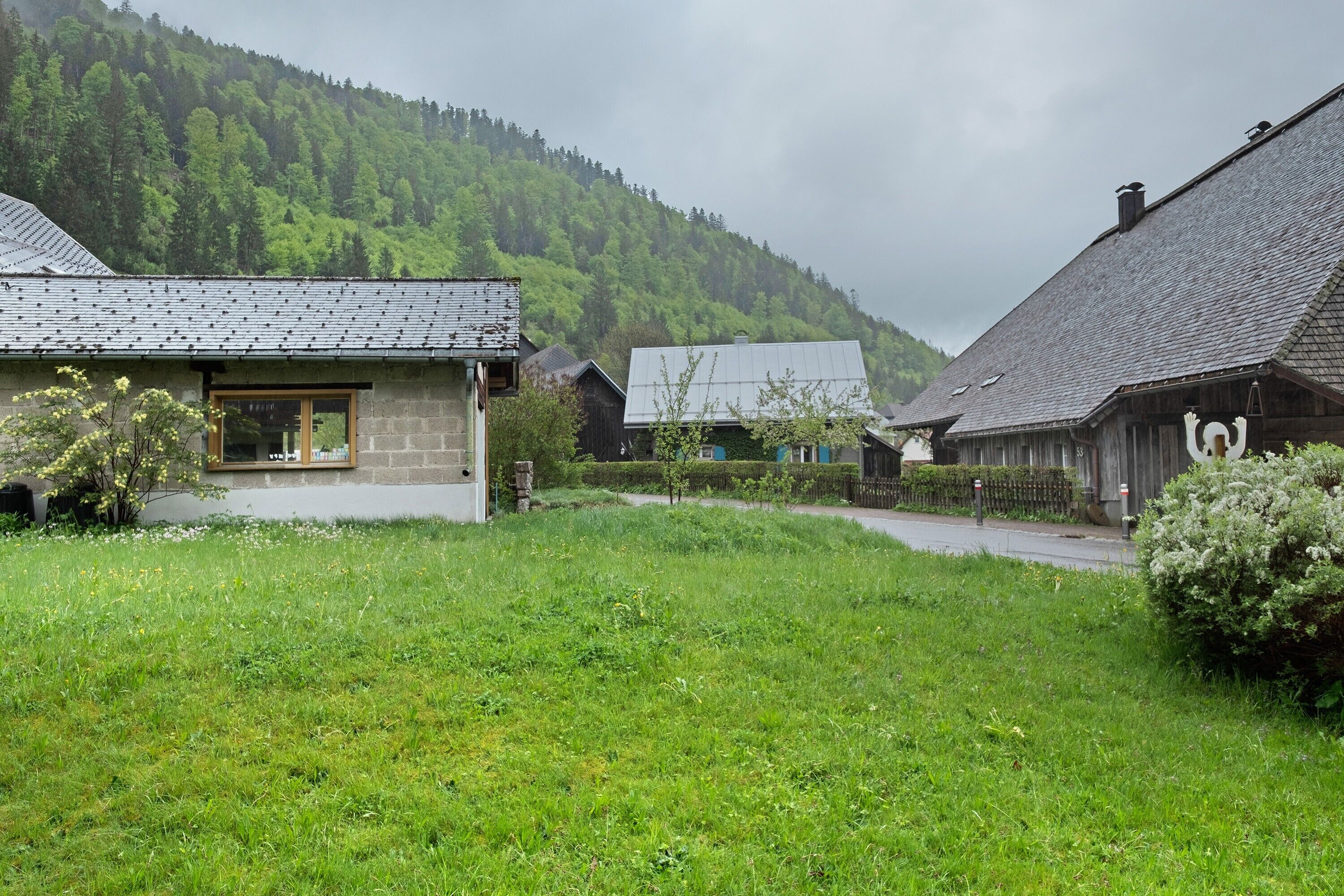 Ein ländliches Dorf mit Steinhäusern, einer grünen Vordergrundfläche und einem bewaldeten Hügel im Hintergrund.