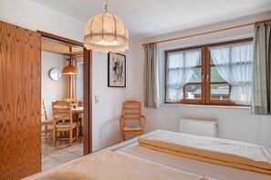 Une chambre à coucher comprenant un lit, une chaise, une table, une lampe, une fenêtre avec des rideaux et un radiateur.