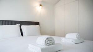 Een net opgemaakt bed met witte linnens en opgerolde handdoeken, een donker hoofdbord en een wandlamp.