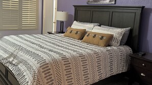 Een netjes opgemaakt bed met een beddegoed met patroon en twee kussens, een nachtkastje met een lamp, en een ingelijst foto aan de muur.