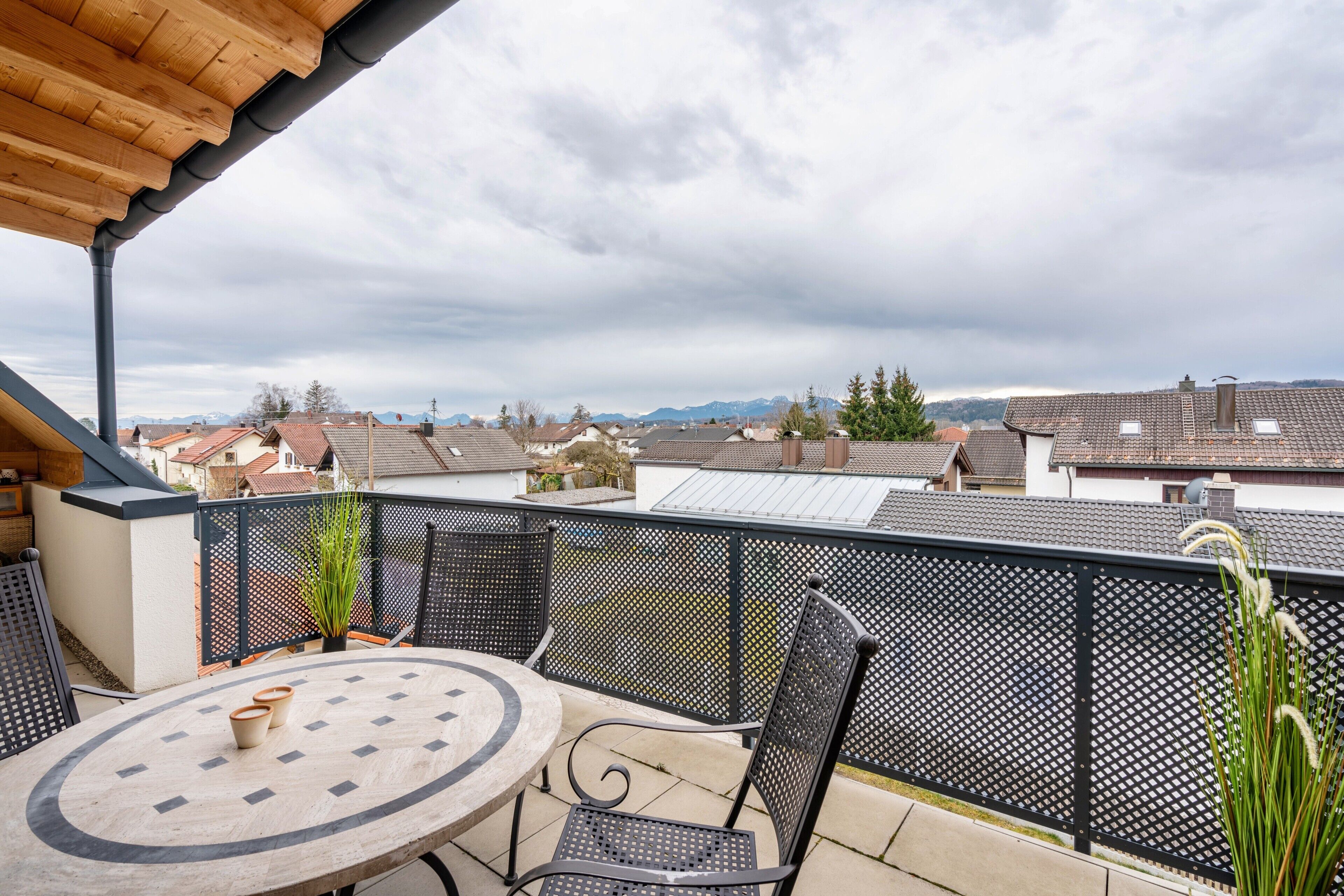 A balcony with a round table and chairs, a view of houses and mountains, and a cloudy sky.