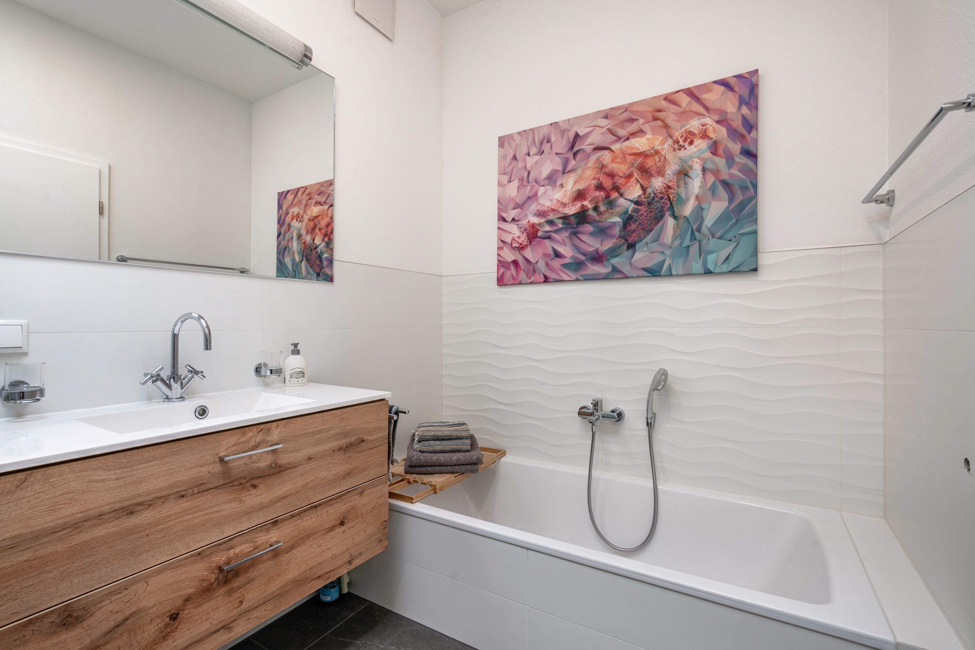 Un baño moderno con un lavabo blanco, tocador de madera, una bañera y dos cuadros abstractos en las paredes.