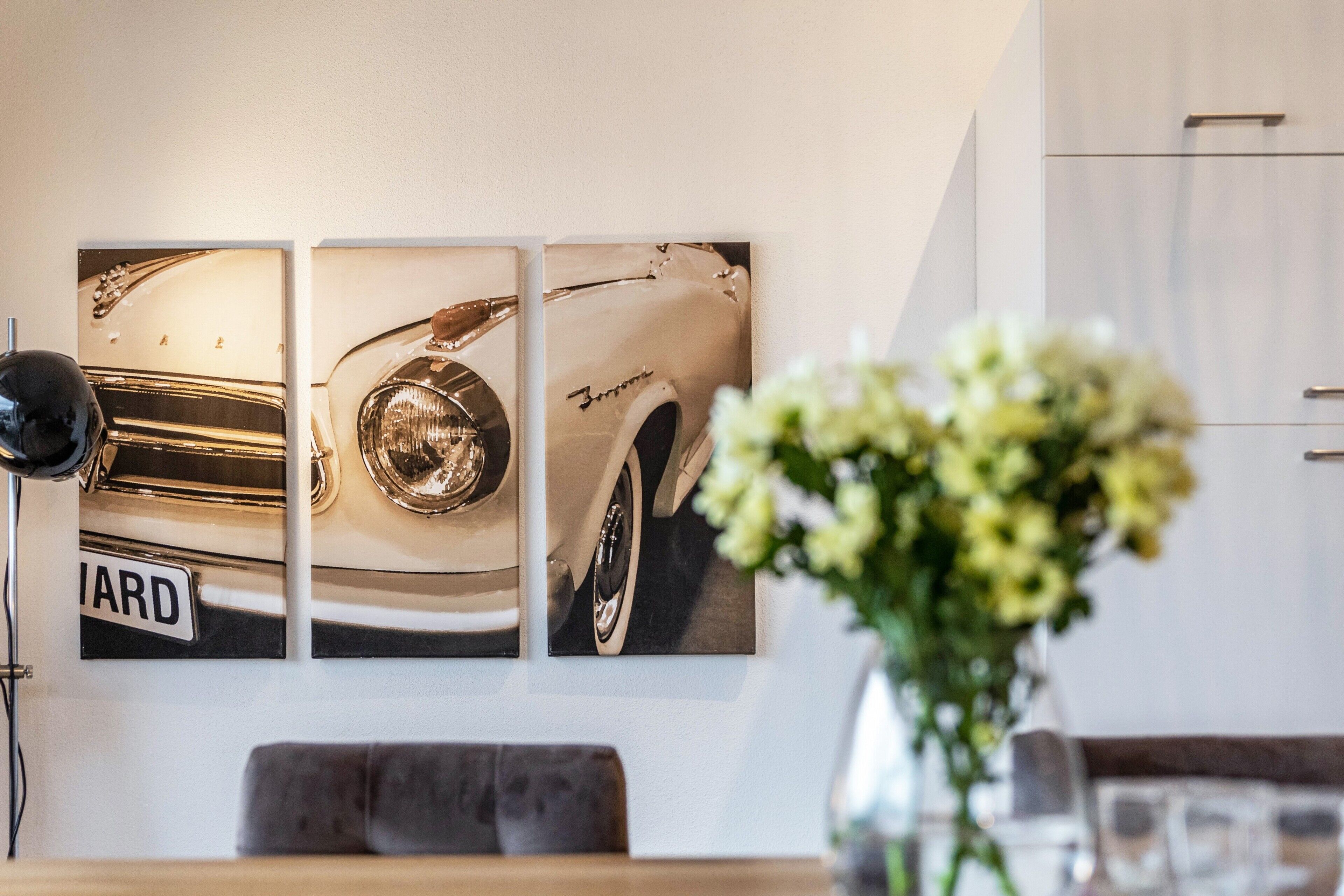 A wall with three framed photographs of a vintage car, a black floor lamp, and a vase with flowers.