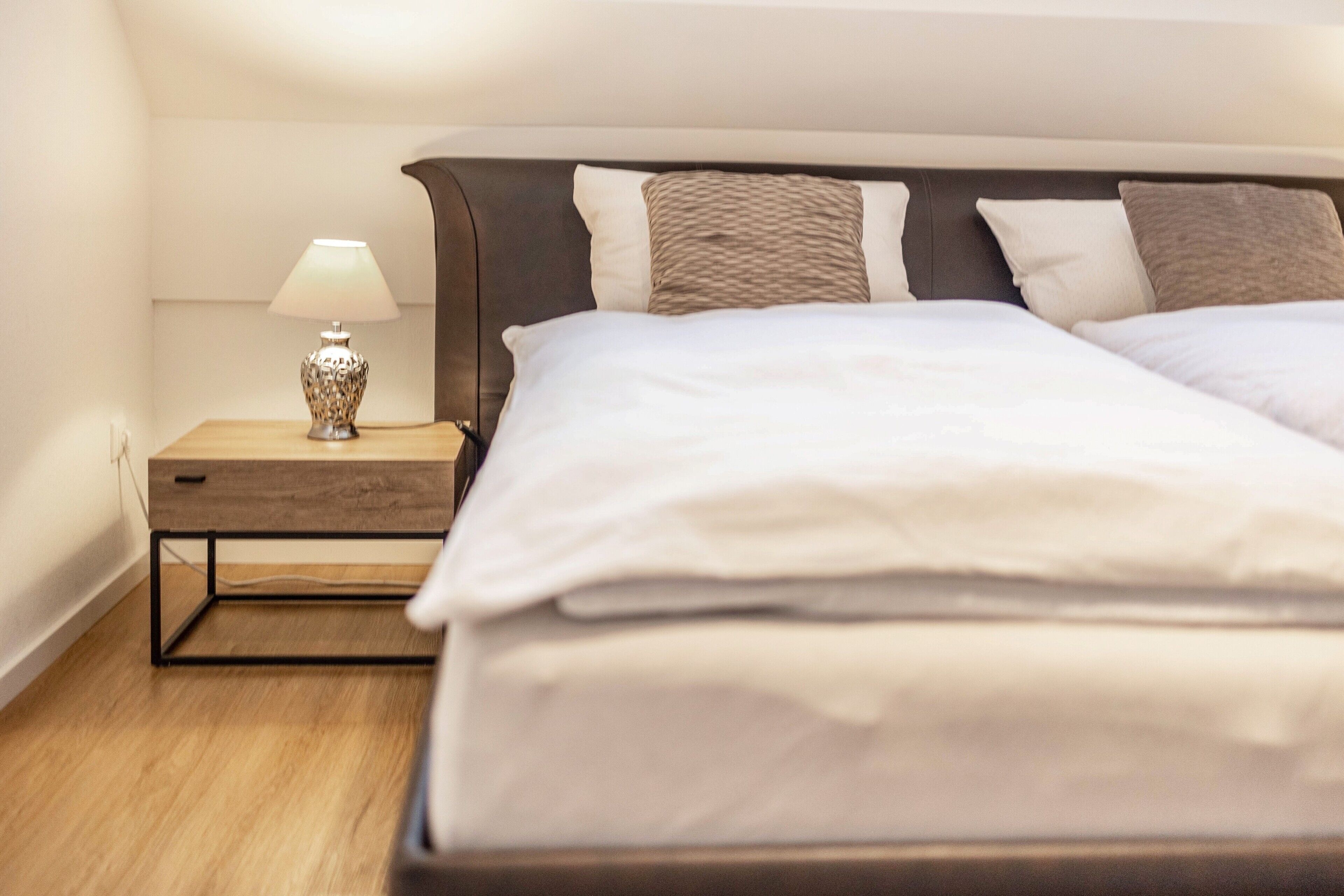 Una cama bien hecha con sábanas blancas, una mesita de noche de madera con una lámpara y un cabecero con varios almohadones.