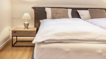 Una cama bien hecha con sábanas blancas, una mesita de noche de madera con una lámpara y un cabecero con varios almohadones.