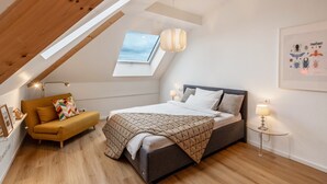 Une chambre avec un plafond en pente, un puits de lumière, un lit, un canapé, une petite table et une lampe fixée au mur.