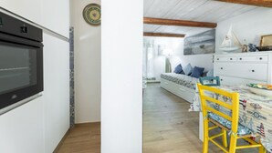 A kitchen with a black oven and a patterned tile border, a dining area with a yellow chair and a table with a nautical-themed tablecloth, and a bedroom with a bed and a painting of a beach scene.
