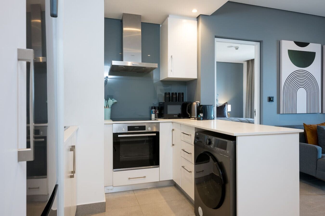 A compact kitchen with a built-in oven, microwave, and dishwasher, white cabinetry, and a grey sofa in the adjacent living area.
