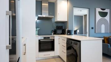 A compact kitchen with a built-in oven, microwave, and dishwasher, white cabinetry, and a grey sofa in the adjacent living area.