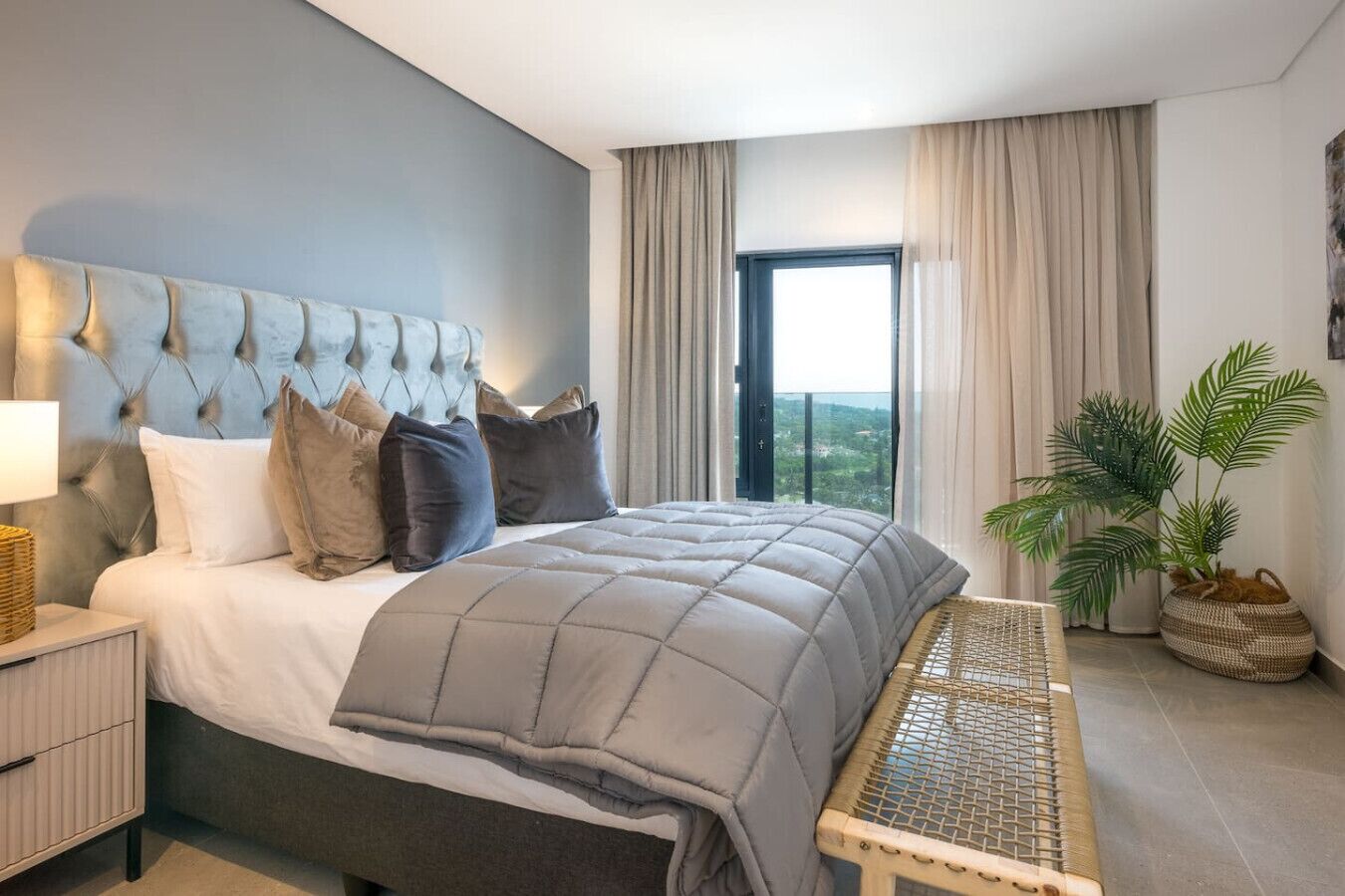 A modern bedroom with a large bed, a headboard with a unique design, a bedside table, a bench, and a view of the outdoors through a window.