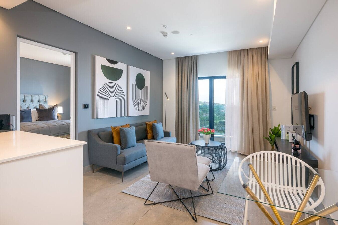 A modern living room with a grey sofa, a white armchair, and a round coffee table. There is a large window with curtains, a flat-screen TV mounted on the wall, and abstract wall art.