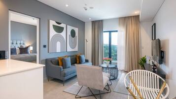 A modern living room with a grey sofa, a white armchair, and a round coffee table. There is a large window with curtains, a flat-screen TV mounted on the wall, and abstract wall art.