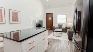 A modern kitchen with a black refrigerator, white cabinets, and a black countertop. There is a television on a stand, a sofa, and a window with blinds. - Sojourn 2 BR Steps to the Beach Sleeps 4 (Virginia Beach)