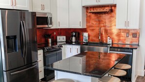 A modern kitchen with stainless steel appliances, white cabinets, a copper backsplash, and a central island with stools. - Sojourn 2 BR Steps to the Beach Sleeps 4 (Virginia Beach)