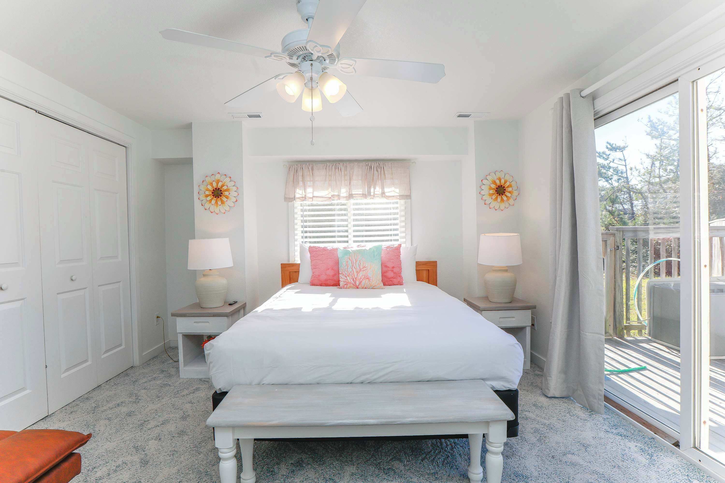 A bedroom with a bed, two bedside tables with lamps, a ceiling fan, a window with blinds, and a view of the outdoors.