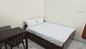 Une petite chambre avec un lit simple, une table et une chaise en bois, une couvre-lit blanche et une porte en bois.